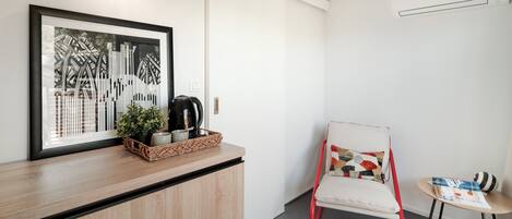 A modern room with a wooden counter, a framed cityscape artwork, a black coffee maker, a red chair, a small round table, and a woven rug.