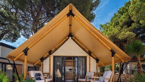 Un espacio al aire libre con toldo, dos sillas de madera y una mesa, rodeado de plantas en macetas y árboles.