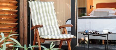 Una silla de madera con un cojín a rayas, una mesita con un cuenco y un libro, y una cama al fondo.