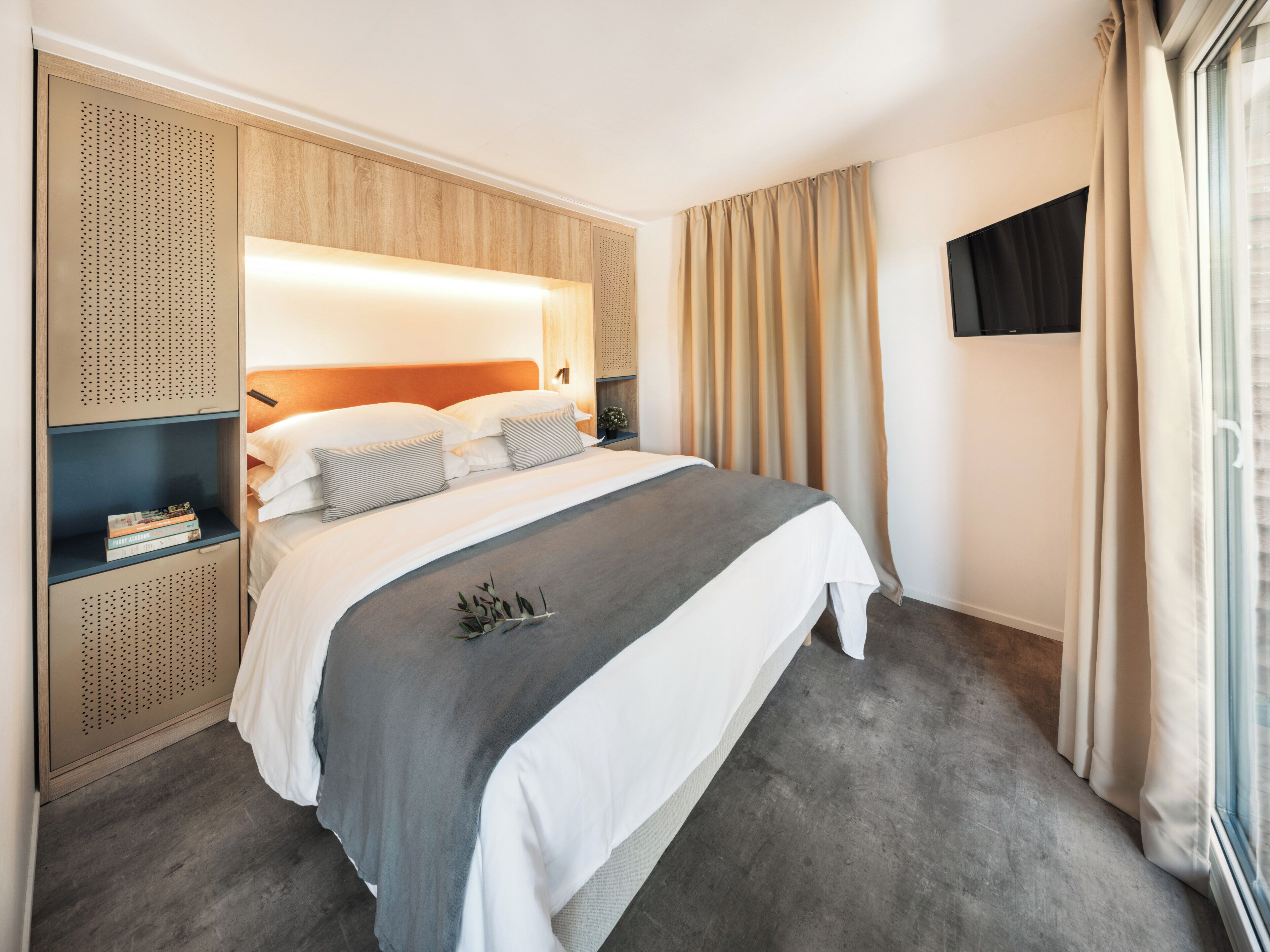 Ein Hotelzimmer mit einem großen Bett, einem hölzernen Kopfteil, einem an der Wand montierten Fernseher und einem Fenster mit Vorhängen.