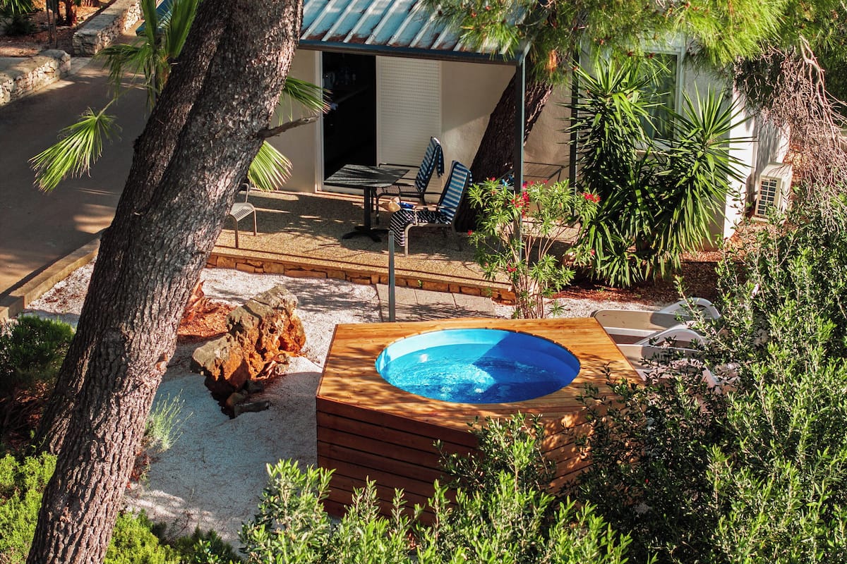A wooden deck with a hot tub, surrounded by lush greenery and a small building with outdoor seating.
