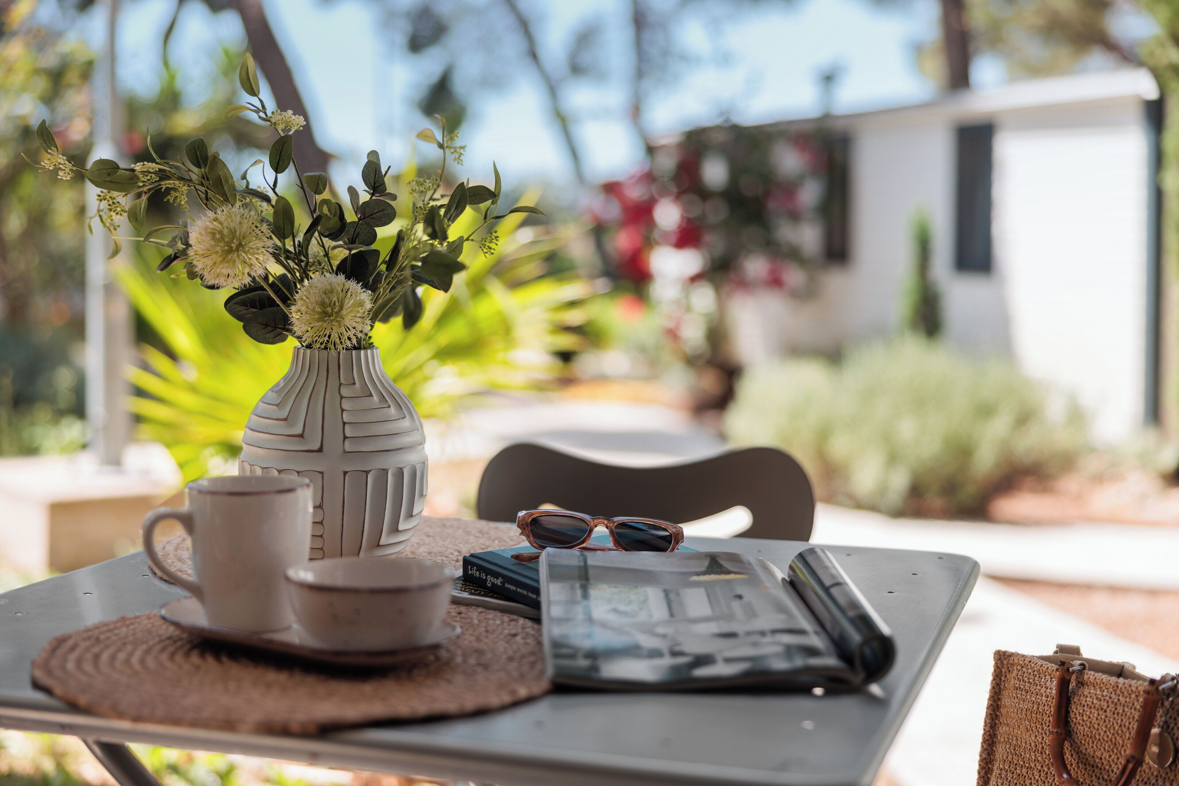 A table with a vase of flowers, a cup, a saucer, a pair of sunglasses, a book, and a smartphone.