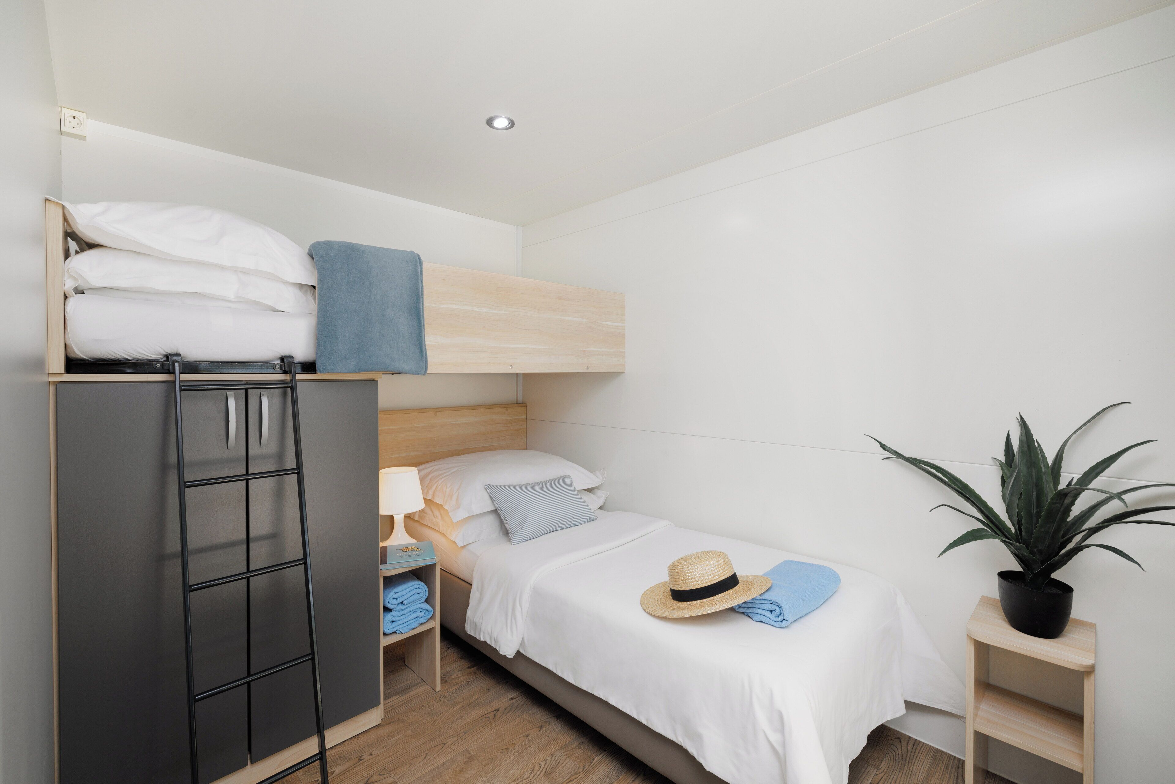 A compact bedroom with a bunk bed, a wardrobe, a bedside table with a lamp, a hat, and a potted plant.