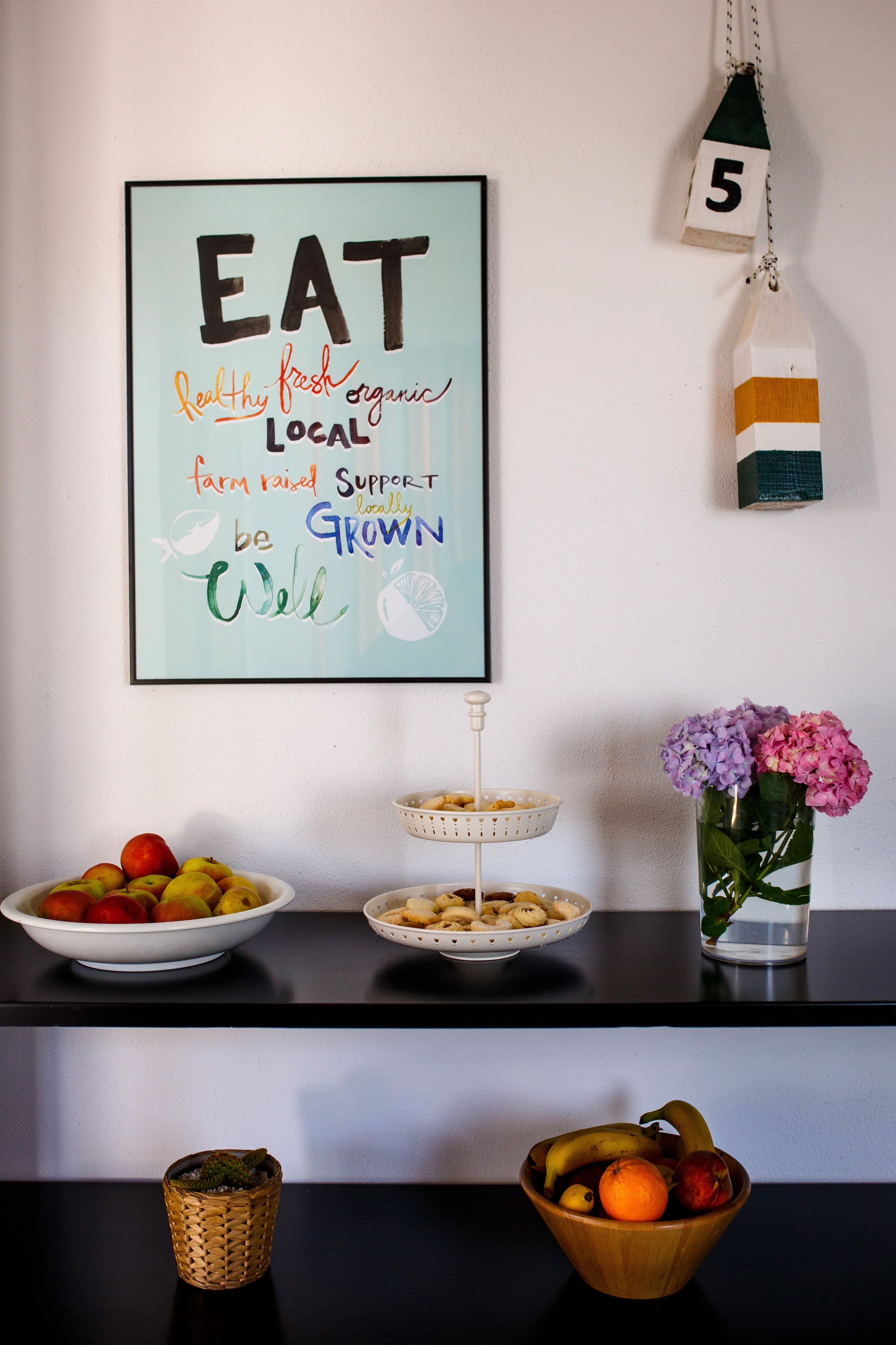 Una encimera de cocina con un frutero, una cesta, un florero y un cartel enmarcado.