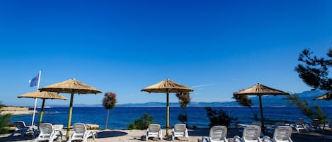 A beach with white lounge chairs and thatched umbrellas, a flag, and a view of the sea and mountains.