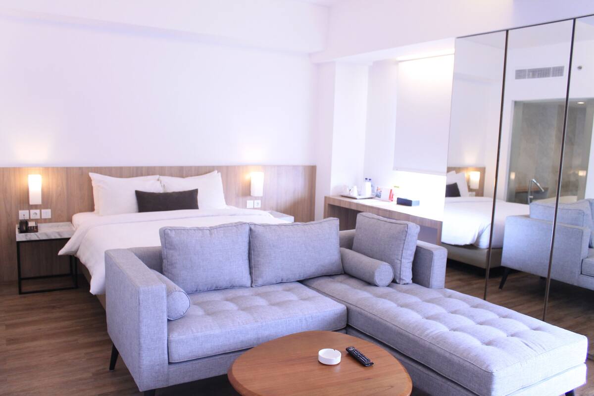 A modern hotel room with a bed, a grey sofa, a wooden coffee table, and a large mirror.
