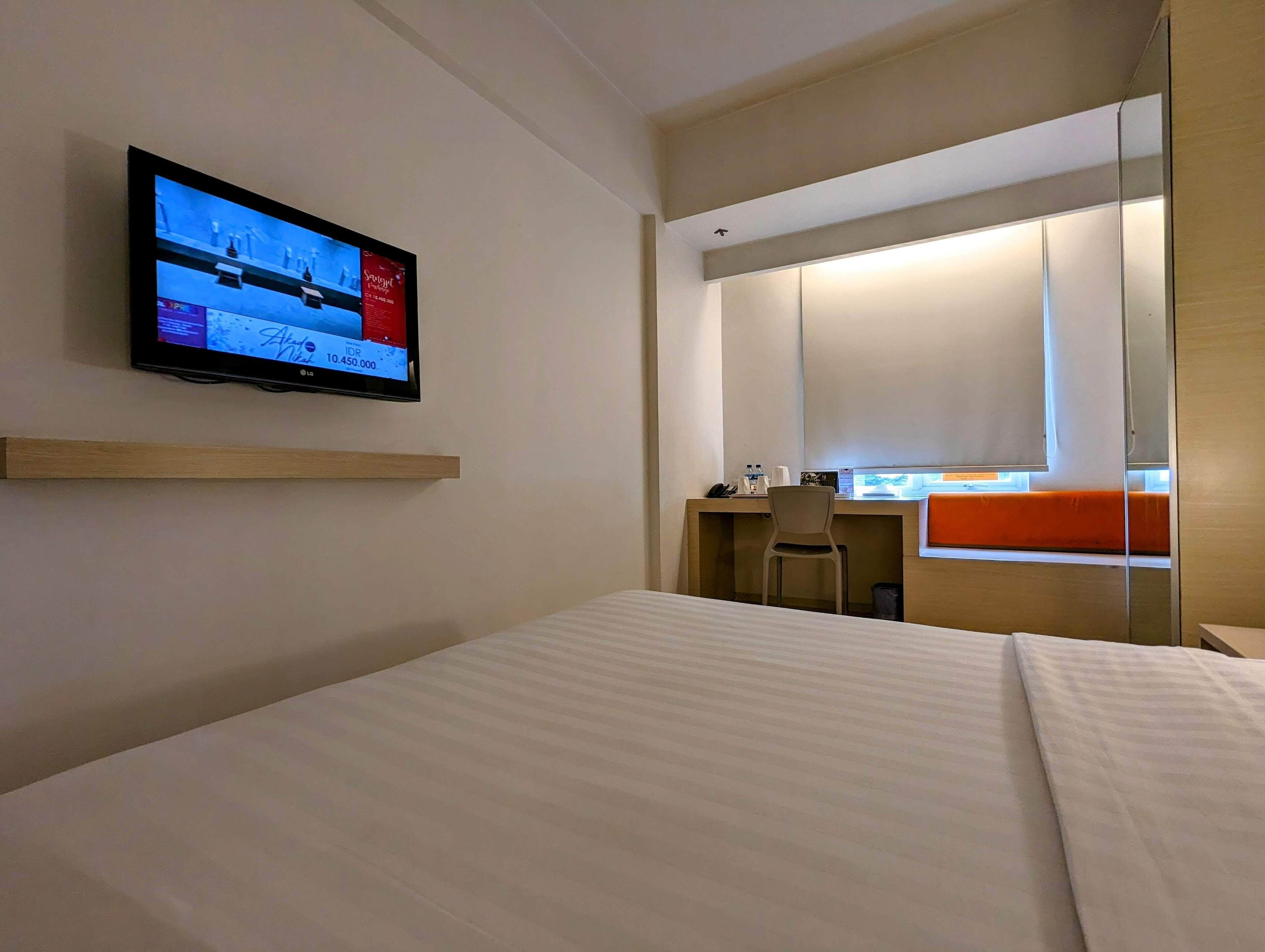 Una camera d'albergo con un letto, una scrivania con sedia, una televisione a parete e una finestra con tende.