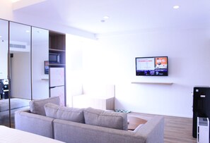 A modern hotel room with a grey sofa, a flat-screen TV mounted on the wall, and a small desk with a computer.