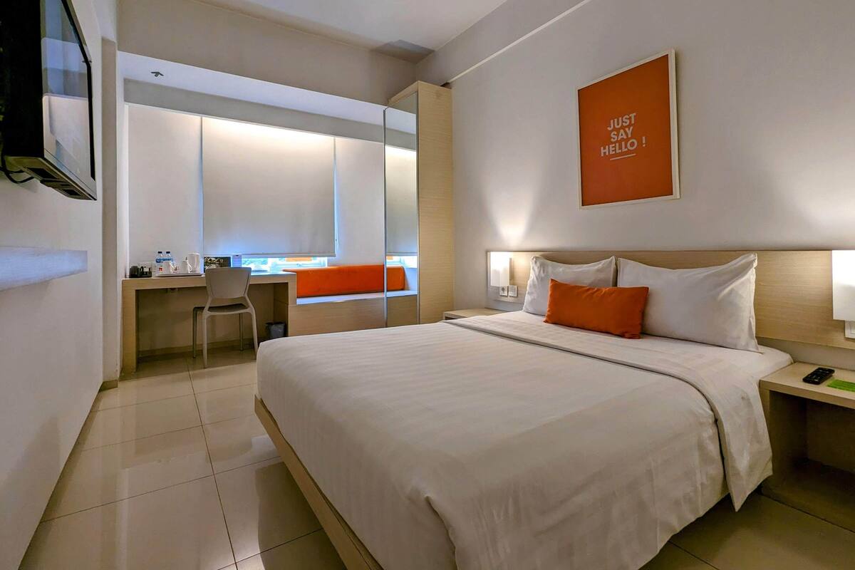 A hotel room with a large bed, a desk, and a chair. There is a mirror on the wall and a sign that says "Just Say Hello".