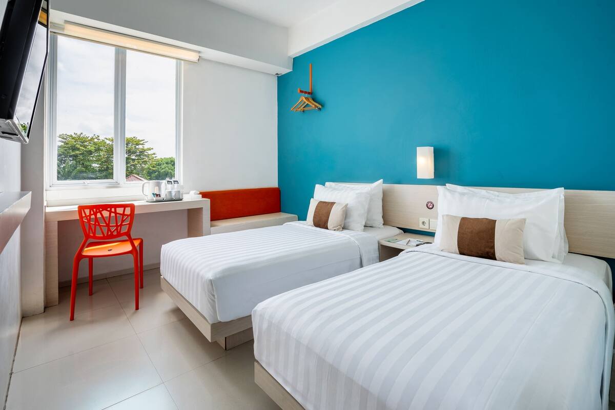Quarto de hotel com duas camas, uma escrivaninha, uma cadeira e uma janela grande.