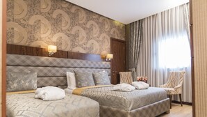 Habitación de hotel con dos camas, cabecera de madera, papel tapiz estampado, una silla y una ventana con cortinas.