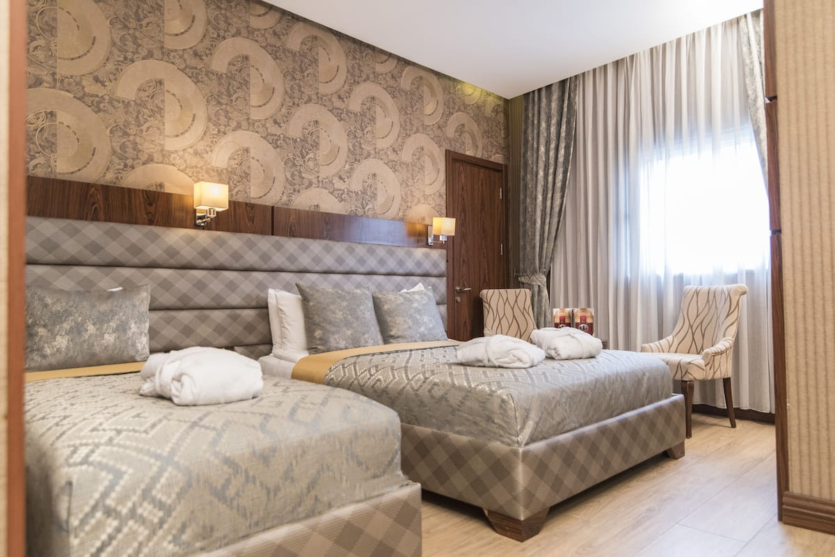 A hotel room with two beds, a wooden headboard, patterned wallpaper, a chair, and a window with curtains.