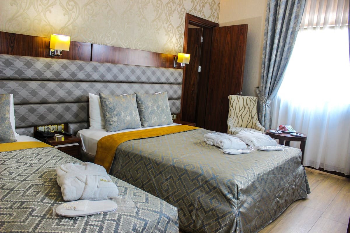 A hotel room with two beds, a wooden headboard, patterned bedding, a nightstand with a lamp, a chair, and a view of the outdoors through a window with curtains.