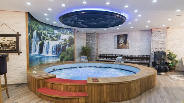 Zona de spa con bañera de hidromasaje, suelo de madera, un mural de una cascada y una estatua de Buda.