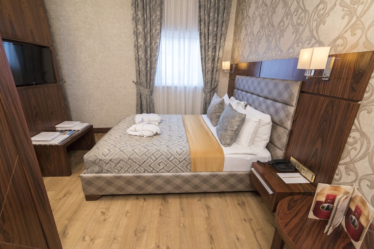 A hotel room with a large bed, a bedside table with a lamp, a TV mounted on the wall, and a wooden wardrobe.