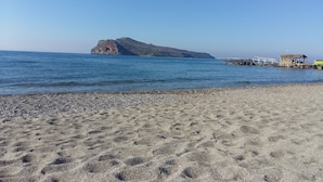 En strand med en klippe i det fjerne, en mole med en lille bygning, og roligt hav.