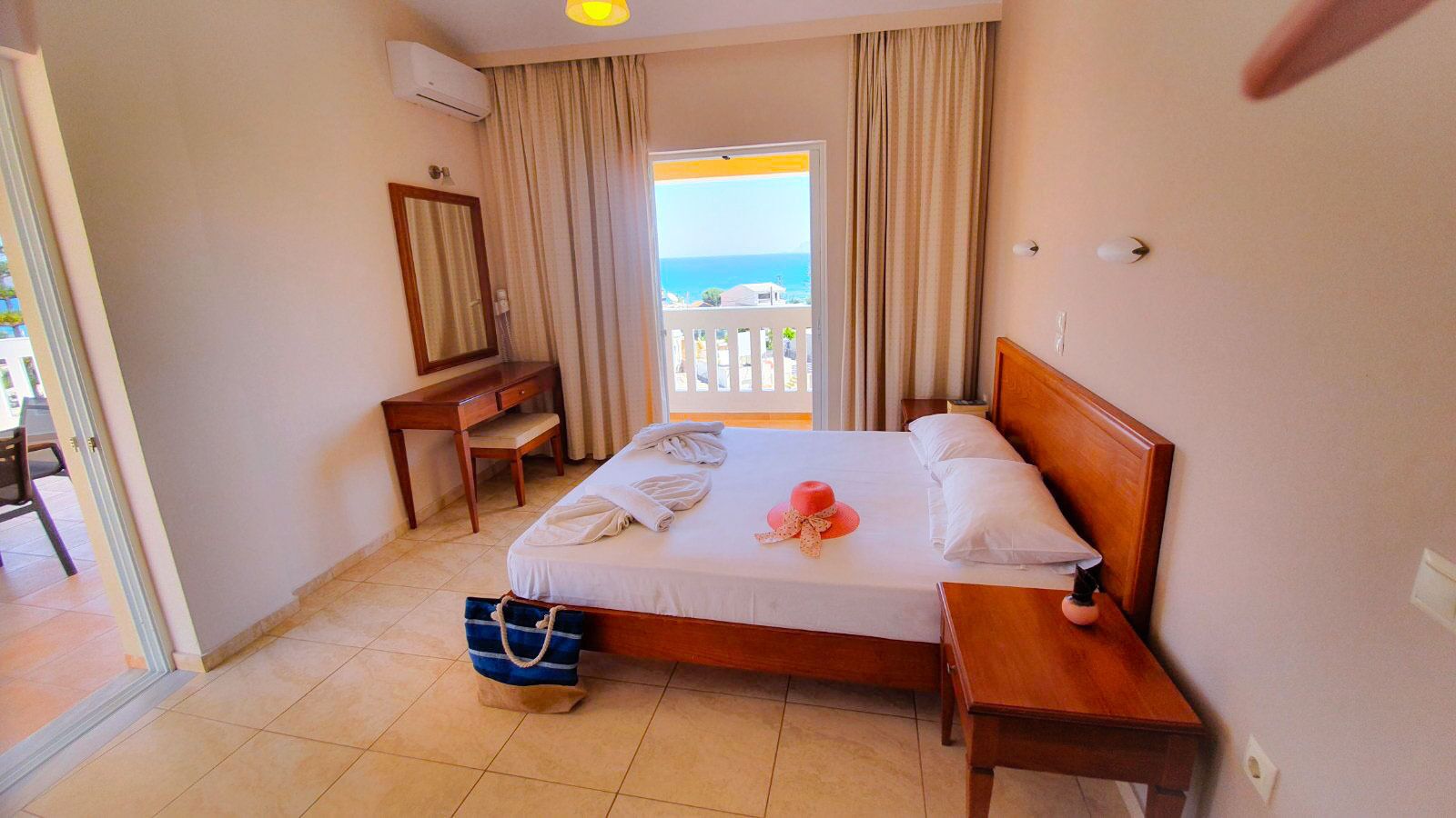 A hotel room with a bed, a desk, a chair, a suitcase, and a view of the sea.