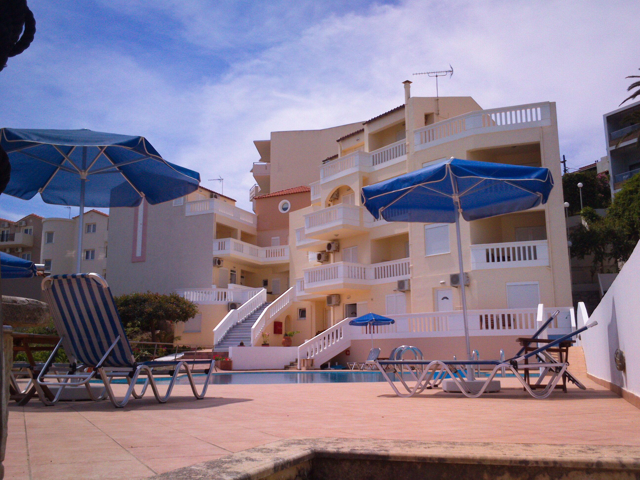 A poolside area with lounge chairs and umbrellas, a staircase leading to a balcony, and a building with multiple balconies and a tiled roof.