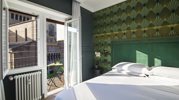 A bedroom with a bed, a radiator, a window with a view of a building, and a patterned wall.
