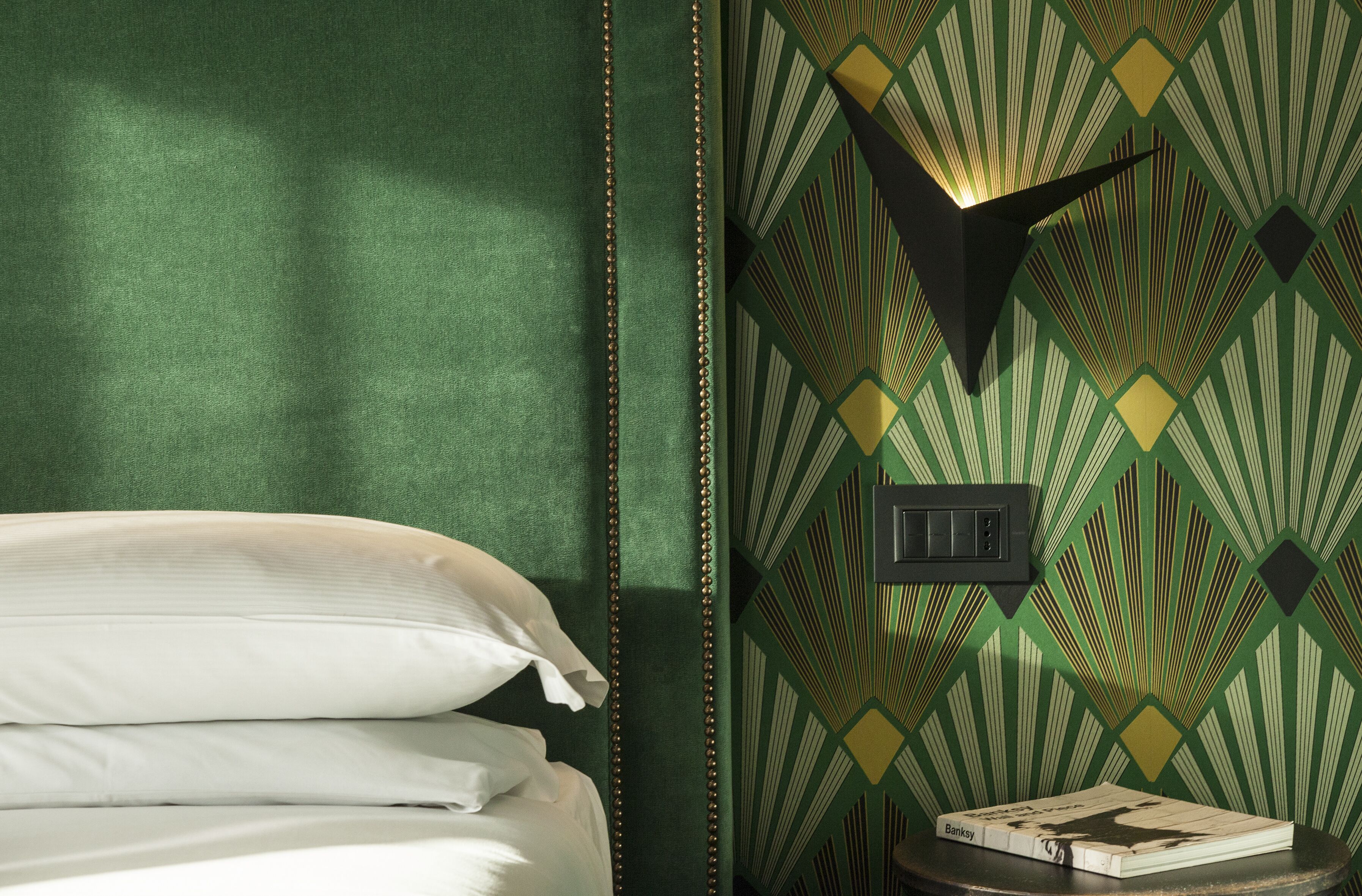 Una cama bien tendida con sábanas blancas, una lámpara de pared y un papel tapiz con diseños geométricos.