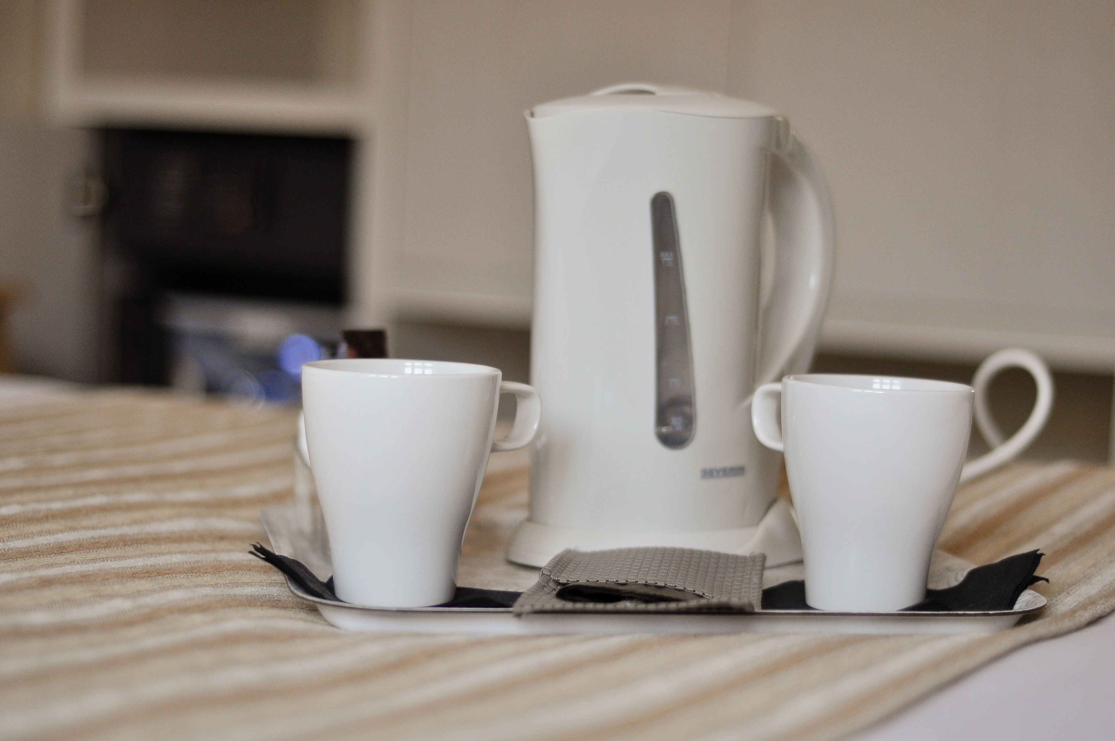 A bed with a striped blanket, a white electric kettle, and two white cups on a tray.