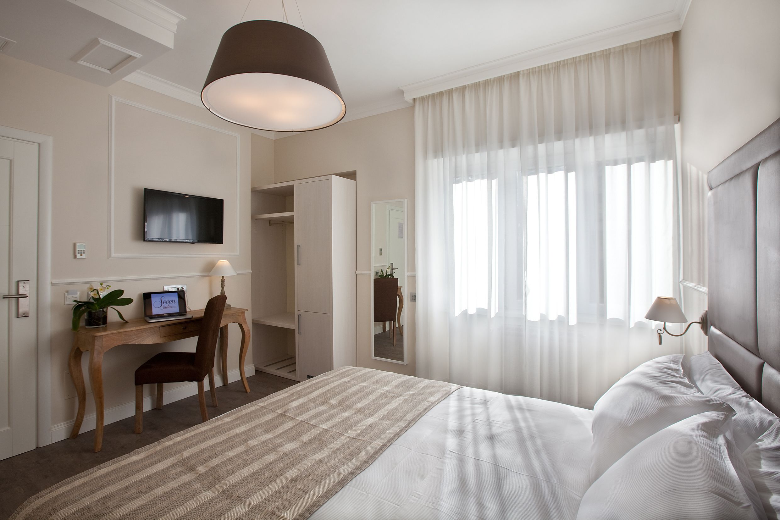 Habitación de hotel con cama, escritorio, computadora, silla, televisión y ventana con cortinas.
