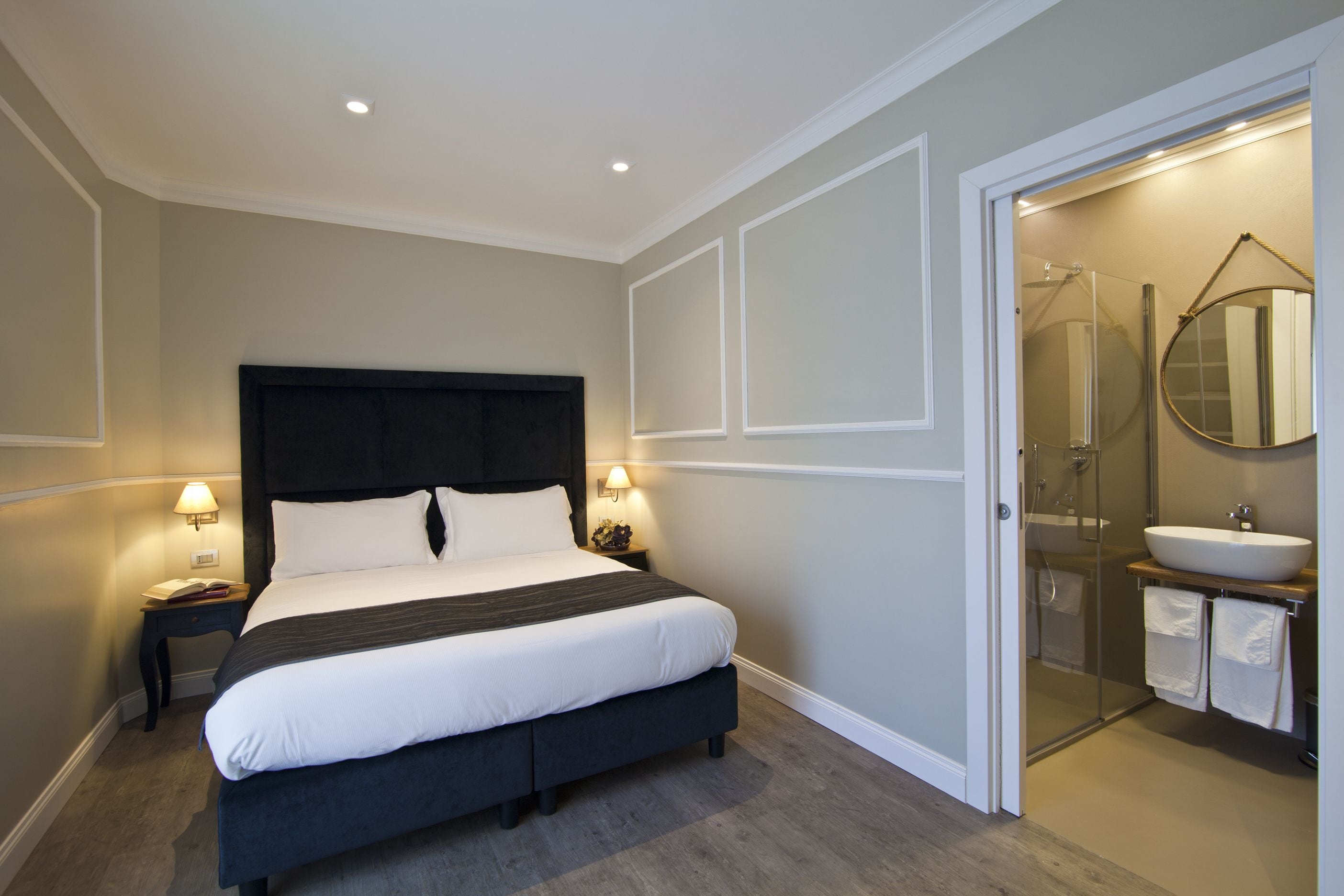 A bedroom with a bed, bedside lamps, a nightstand, and a bathroom with a sink and mirror.