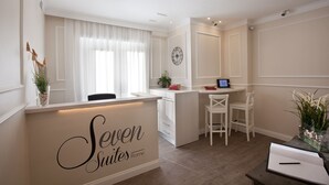 A reception desk with a sign reading "Seven Suites Rome". There is a window with sheer curtains, a clock on the wall, and a small desk with a laptop and a plant.