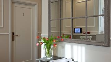 Una habitación con una puerta que tiene el número 1, un ventanal con un espejo que refleja otra habitación, y un florero con tulipanes sobre un escritorio.