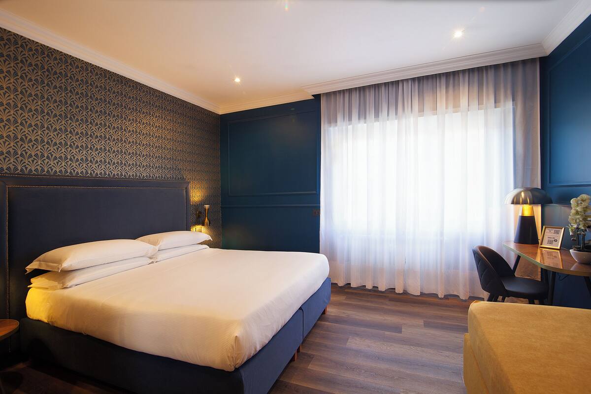 A modern hotel room with a large bed, a desk with a lamp, and a view of the outdoors through sheer curtains.