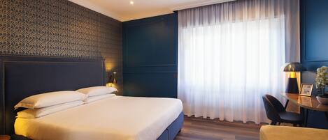 A modern hotel room with a large bed, a desk with a lamp, and a view of the outdoors through sheer curtains.