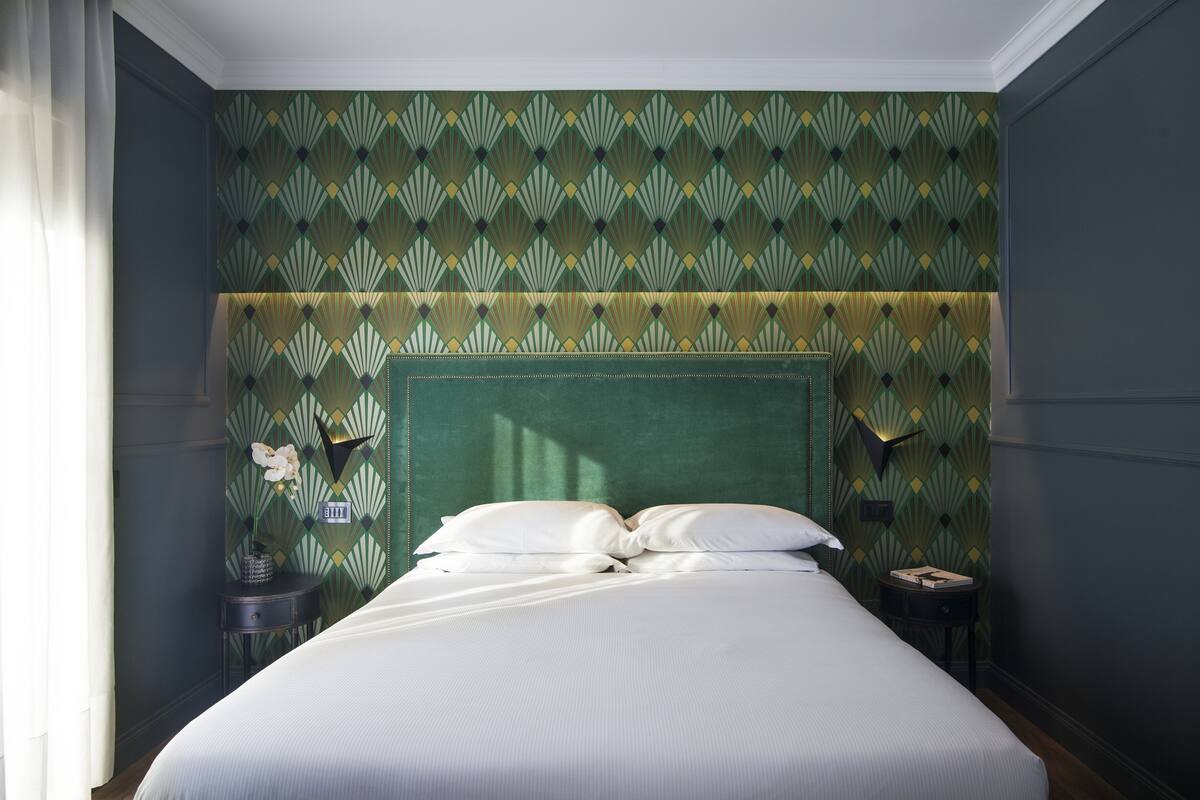 A neatly made bed with white linens in a room with green patterned wallpaper and a green upholstered headboard.