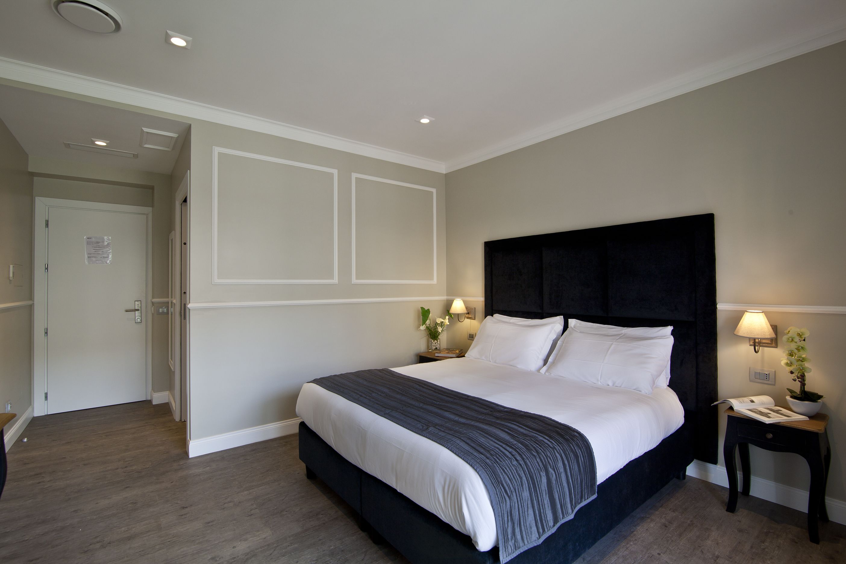 A neatly made bed with a dark headboard, two bedside lamps, and a small table with a book and a plant.