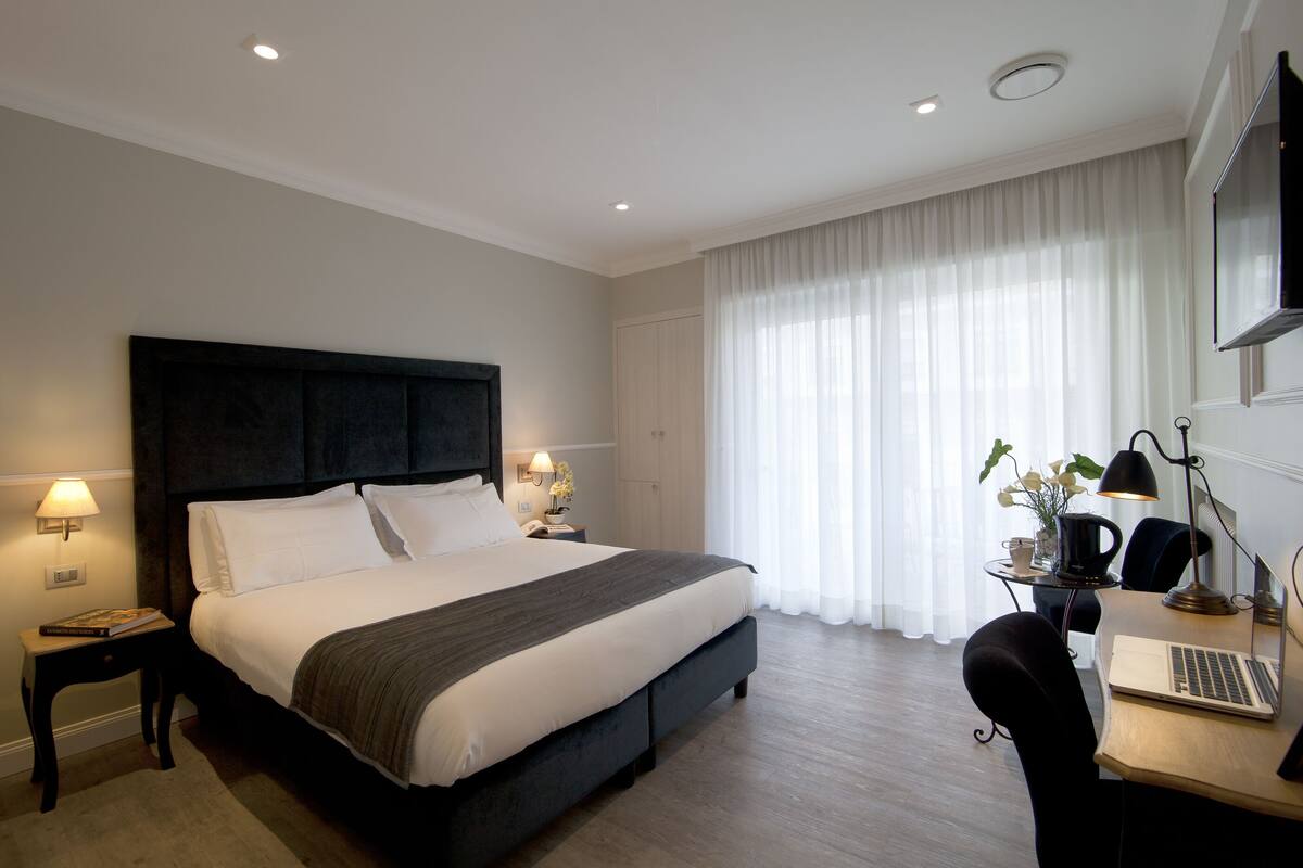 A modern hotel room with a large bed, bedside lamps, a desk with a laptop, and a large window with sheer curtains.