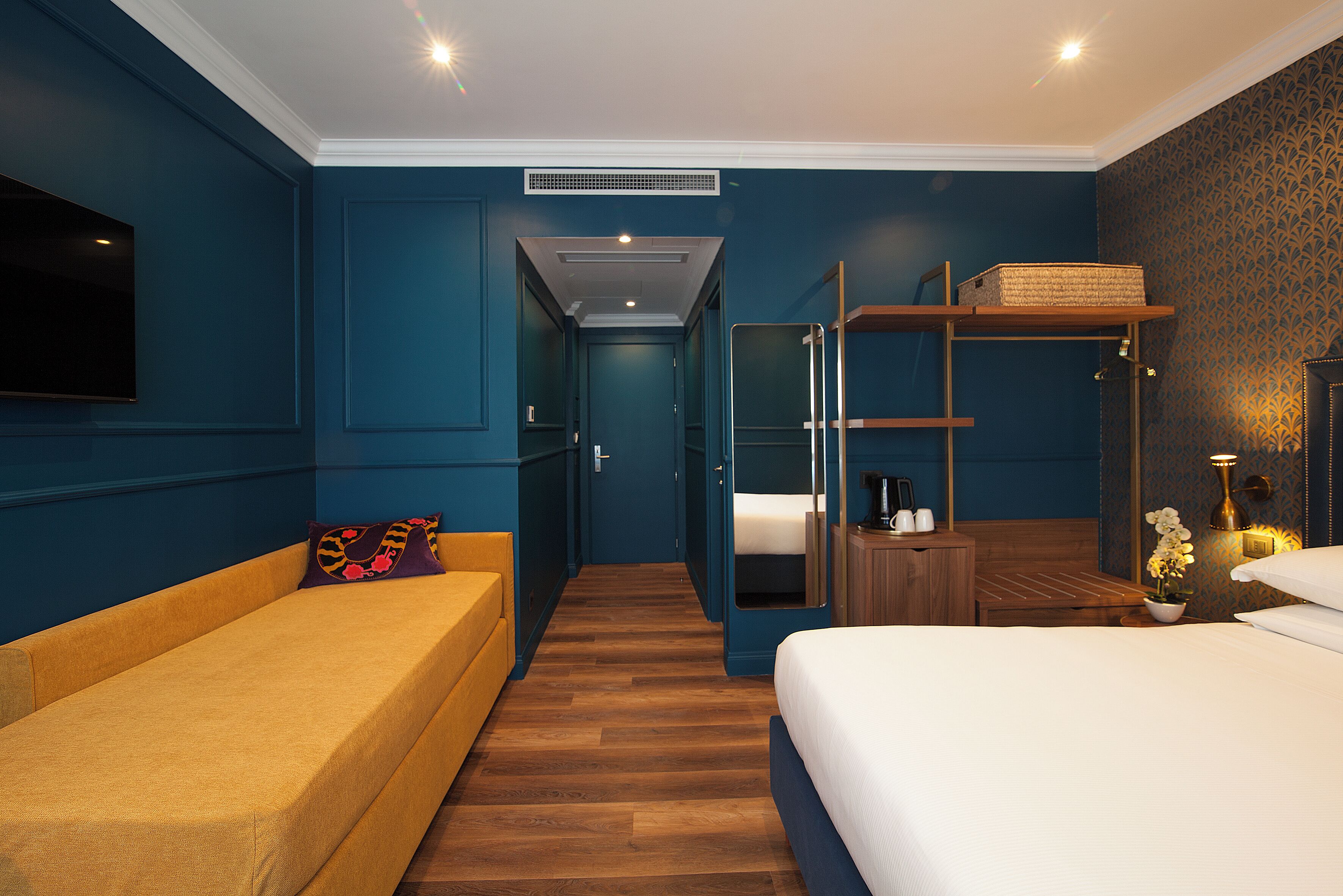Habitación de hotel con cama, sofá, televisor, estante con cesta y mesita de noche con lámpara y flores.