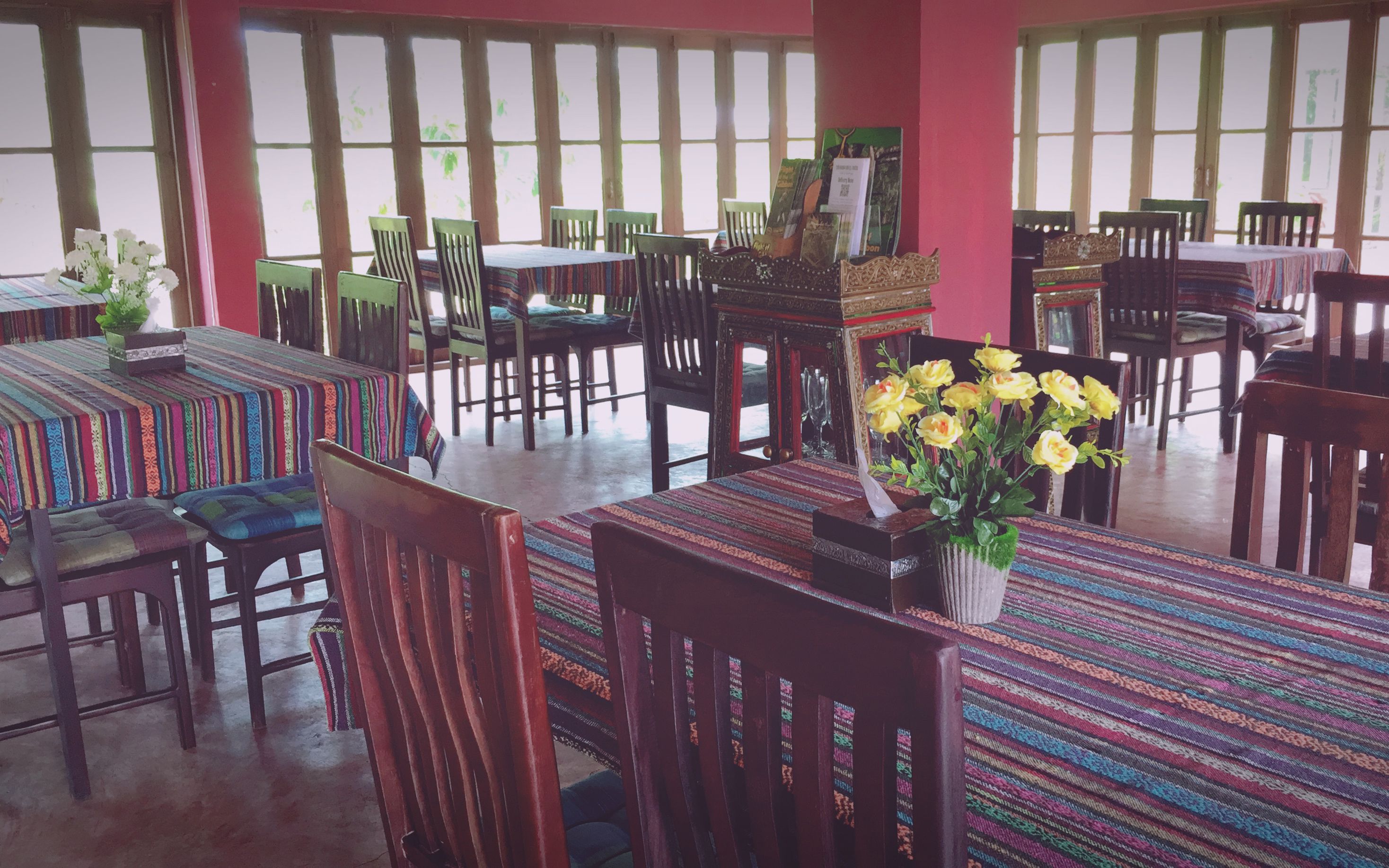 A dining area with tables covered in colorful tablecloths, wooden chairs, and a vase of yellow flowers.