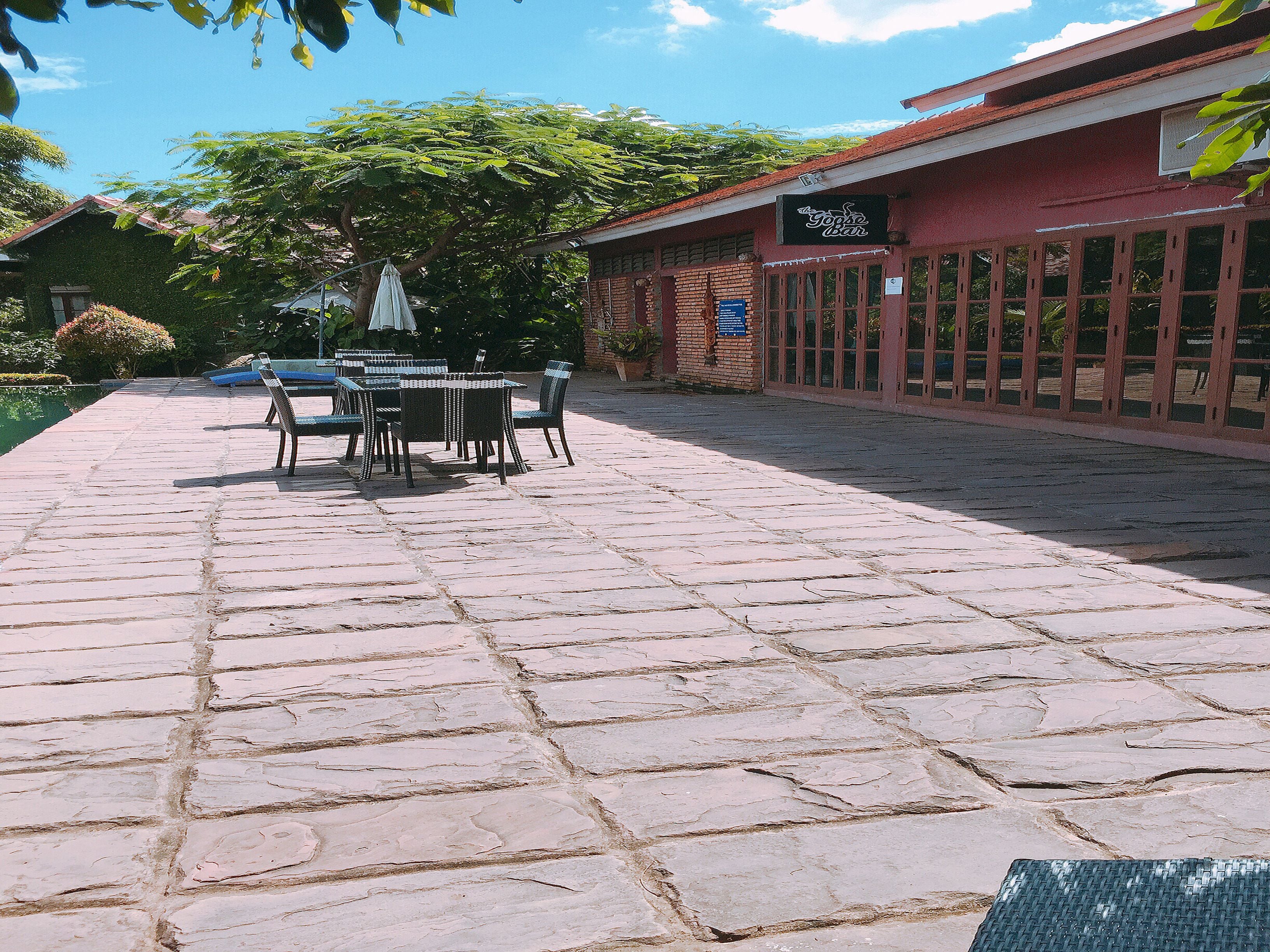 A paved outdoor area with a table and chairs, a building with a sign, and a tree in the background.