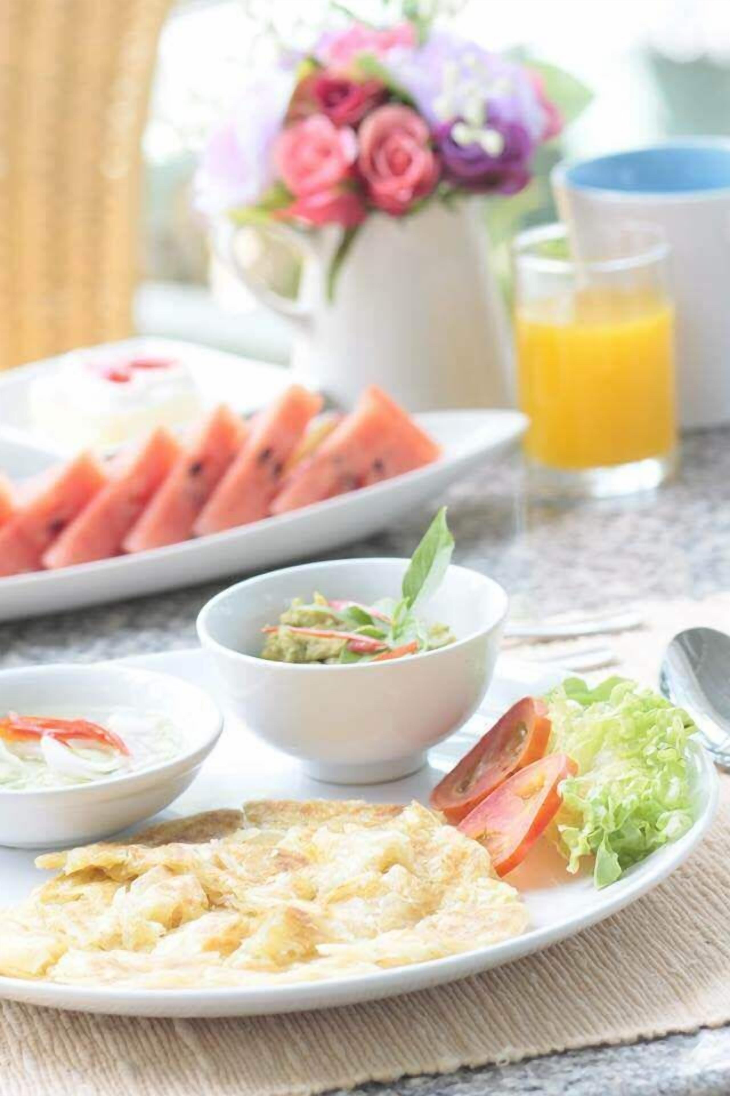 Une table dressée pour le petit-déjeuner avec des tranches de pastèque, un bol de soupe, une assiette d’œufs brouillés et un verre de jus d’orange.