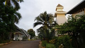 A residential street with houses, a prominent white building with a pointed roof, and lush greenery including palm trees. - A Little Villa (Krabi)