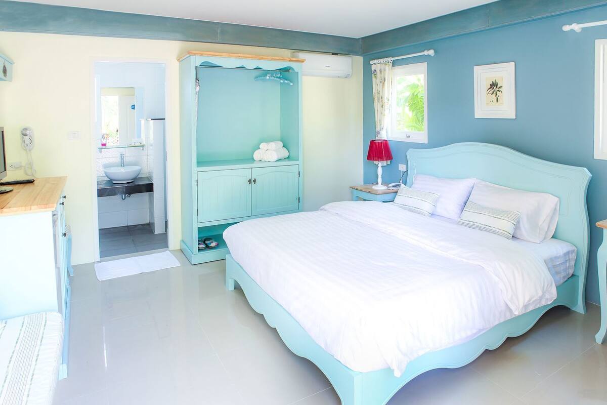 A bedroom with a bed, a nightstand, a red lamp, a window with curtains, and a bathroom visible through an open door.