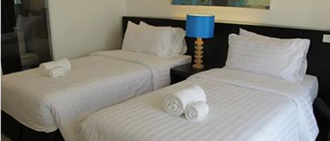 Two single beds with white linens and rolled towels, a nightstand with a blue lamp, and a colorful abstract painting on the wall.