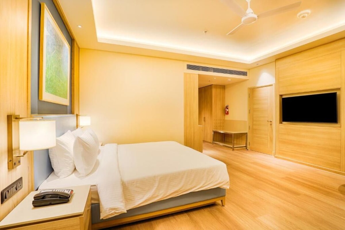 A hotel room with a bed, bedside table, lamp, ceiling fan, TV, and wooden walls.