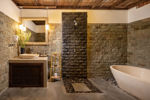 A modern bathroom with a stone wall, a freestanding bathtub, a sink with a mirror, and a wooden shelf with toiletries.