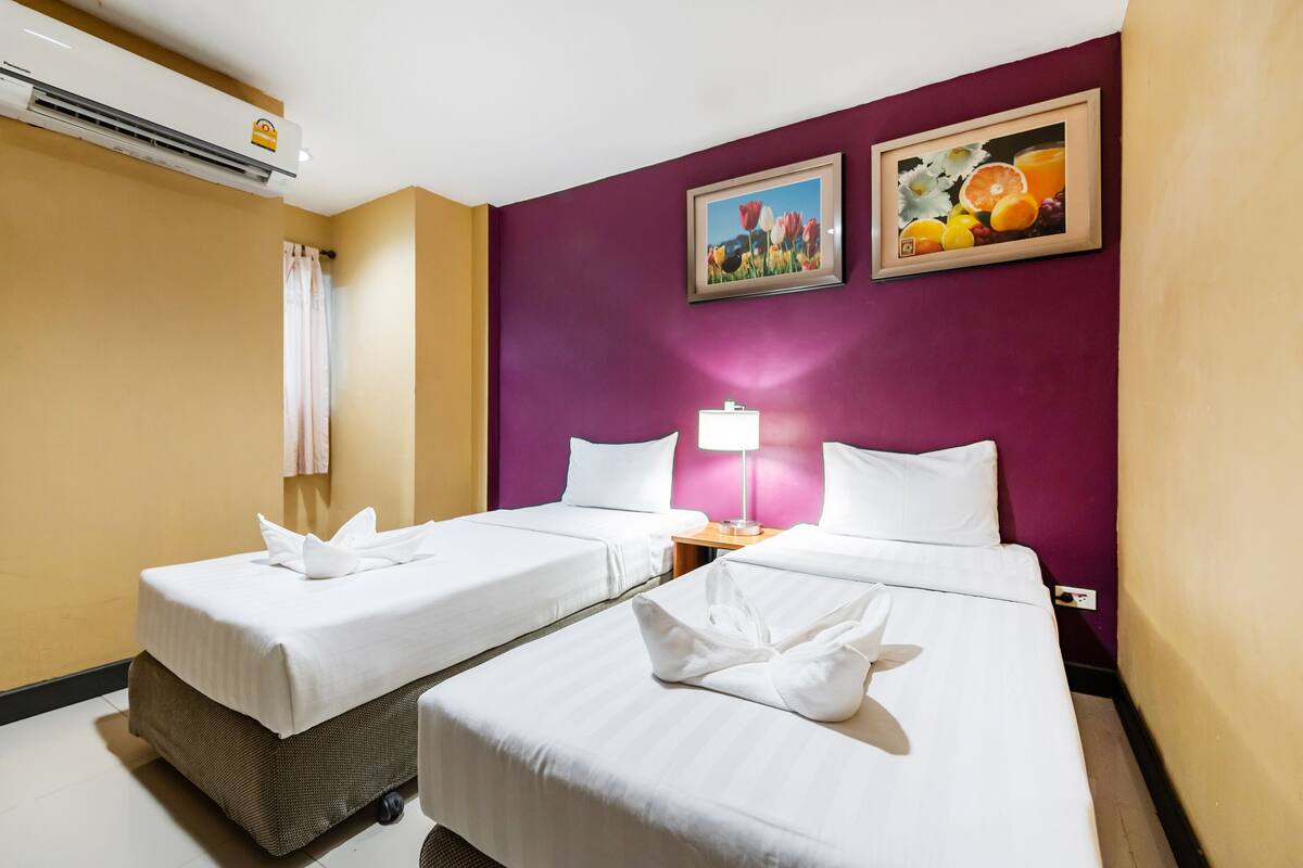 Une chambre d’hôtel avec deux lits, un climatiseur fixé au mur, un tableau représentant des fleurs et une lampe.