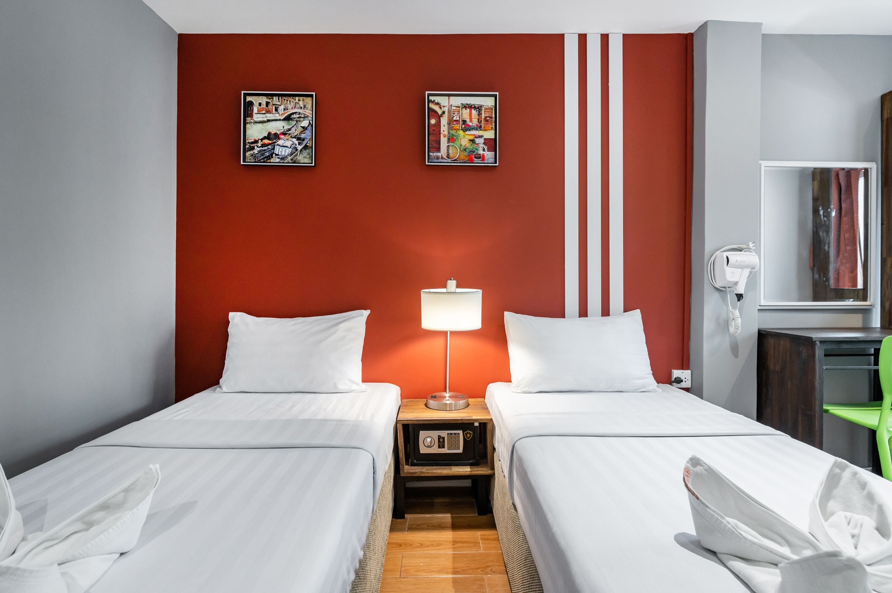 A hotel room with two beds, a red accent wall, a lamp, a mirror, and artwork.