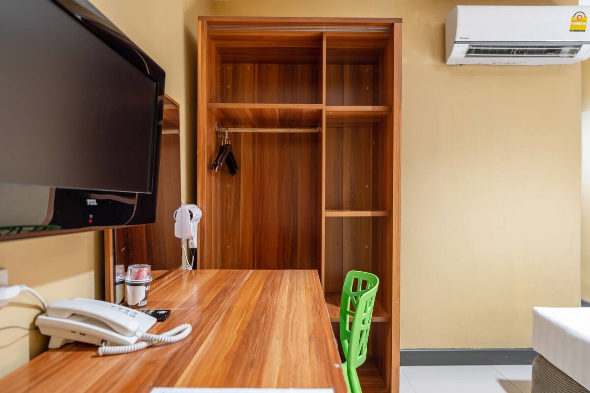 Une chambre d’hôtel avec un bureau en bois, un téléphone, une chaise verte, un téléviseur et une armoire contenant des vêtements suspendus.