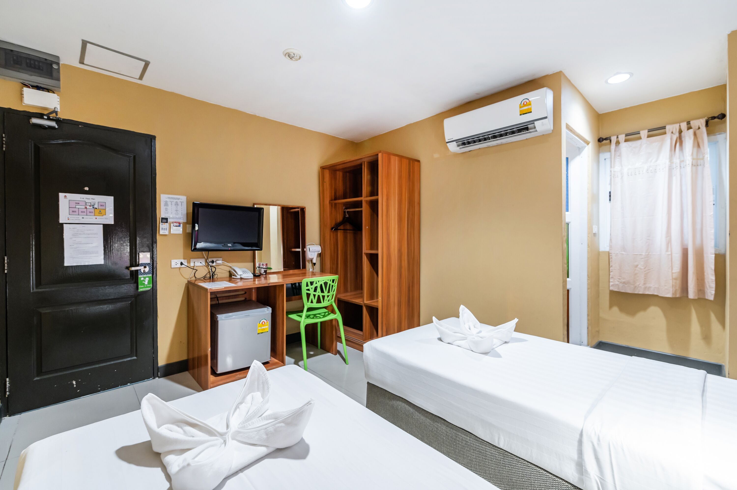 Une chambre d’hôtel avec deux lits, un bureau avec un ordinateur, un petit réfrigérateur, une chaise verte et une armoire.