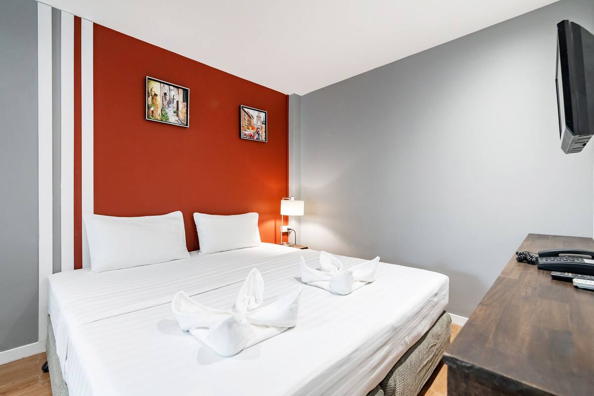 Une chambre d’hôtel avec un lit bien fait, deux tableaux encadrés au mur, une lampe de chevet, un bureau avec un téléphone et un téléviseur fixé au mur.