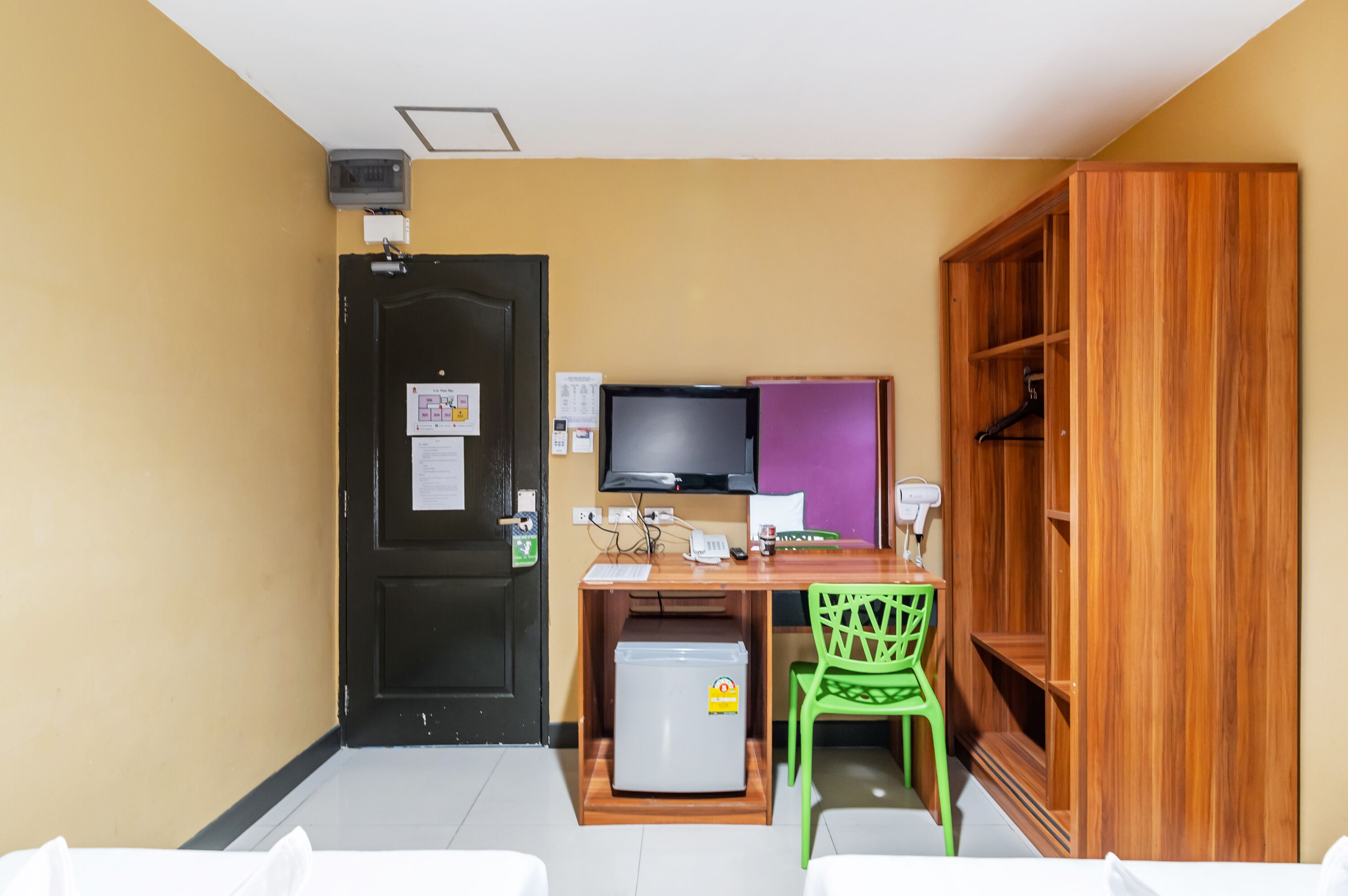 A compact room with a wooden desk, a television, a small refrigerator, a green chair, and a wardrobe.