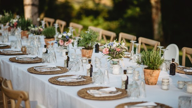 A long table set for a meal outdoors with white tablecloths, woven placemats, and potted plants. - Agriturismo San Galgano (Chiusdino)